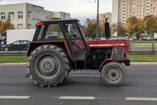 Czerwony Ursus C385 z kabiną jedzie ulicą, obok budynków mieszkalnych i drzew.