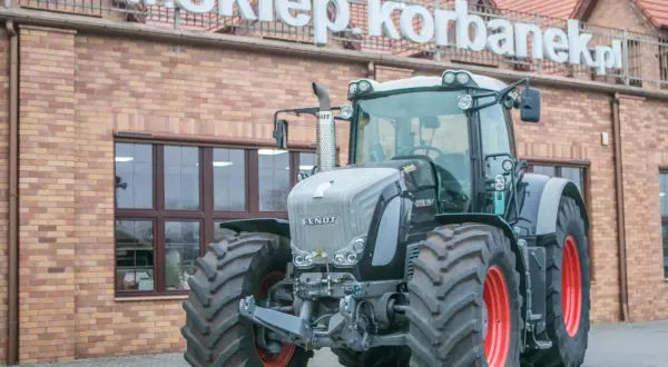 Potężny traktor Fendt 936 z czerwonymi felgami stoi przed budynkiem z napisem "www.sklepkorbanek.pl".