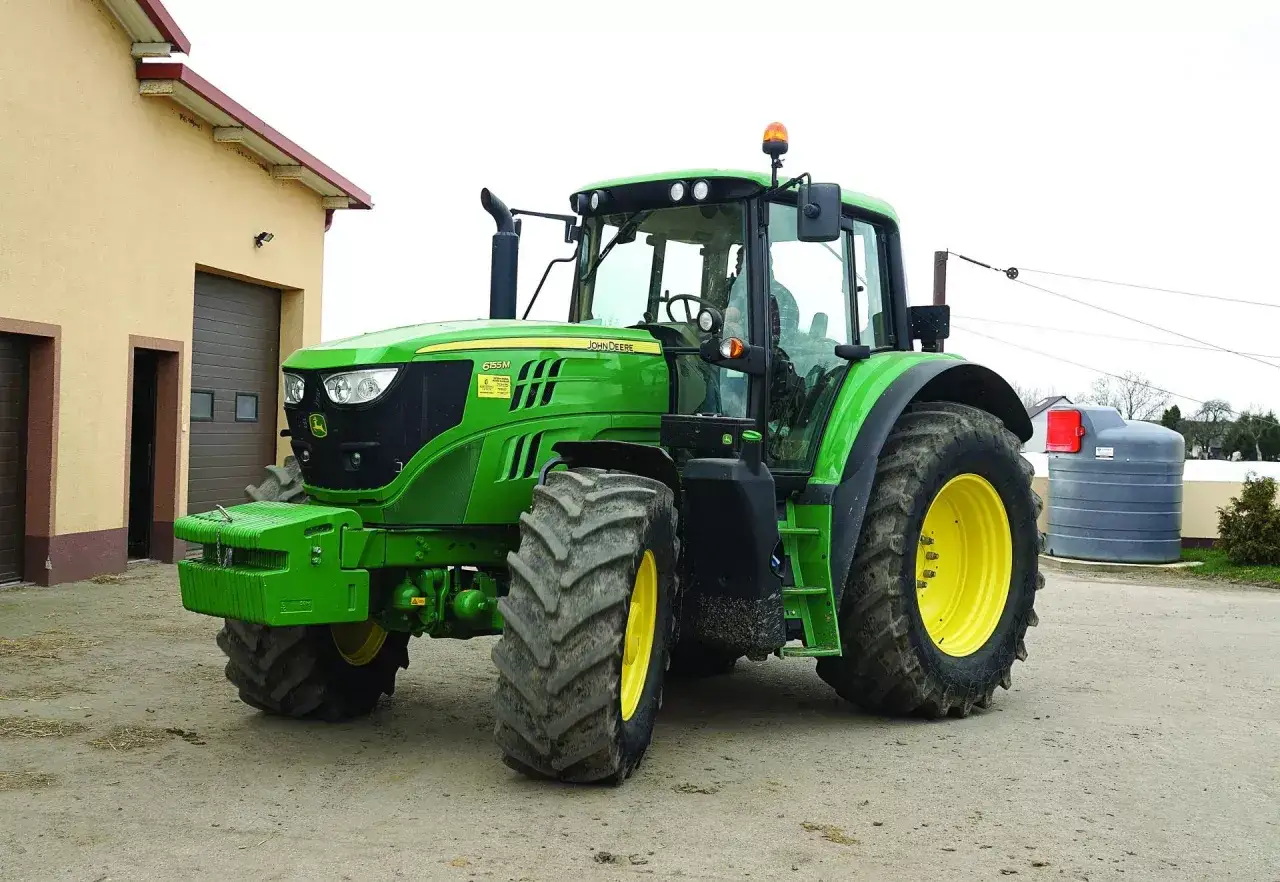 Zielony traktor John Deere, jeden z popularnych marek traktorów, stoi przed budynkiem gospodarczym.